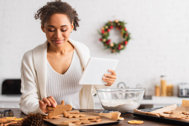 Mujer afroamericana sosteniendo tableta digital cerca de pan de jengibre y harina en la cocina  - Foto, imagen