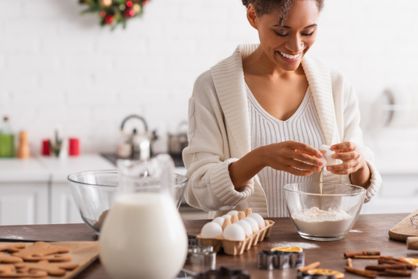 幸せなアフリカ系アメリカ人の女性は、ぼやけたクッキーとシナモンスティックの近くに小麦粉に卵を注ぐ  - 写真・画像