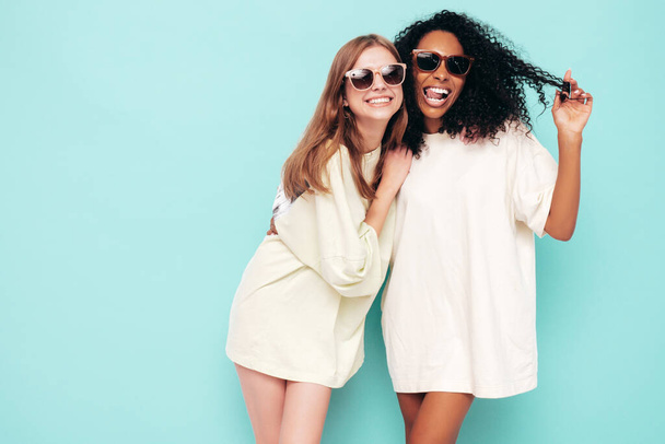Two young beautiful smiling international hipster female in trendy summer clothes. Sexy carefree women posing near blue wall in studio. Positive models having fun in sunglasses. Concept of friendship - Foto, afbeelding