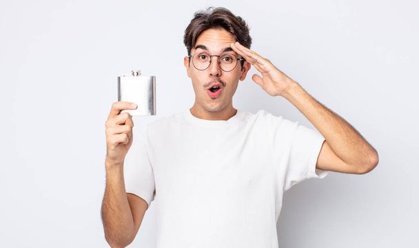 young hispanic man looking happy, astonished and surprised. alcohol flask concept - Zdjęcie, obraz