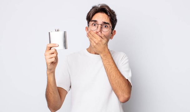 young hispanic man covering mouth with hands with a shocked. alcohol flask concept - Foto, Imagen