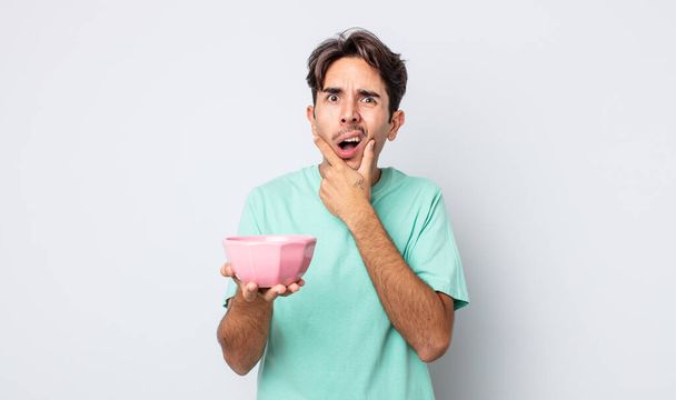 young hispanic man with mouth and eyes wide open and hand on chin. empty bowl concept - Фото, изображение