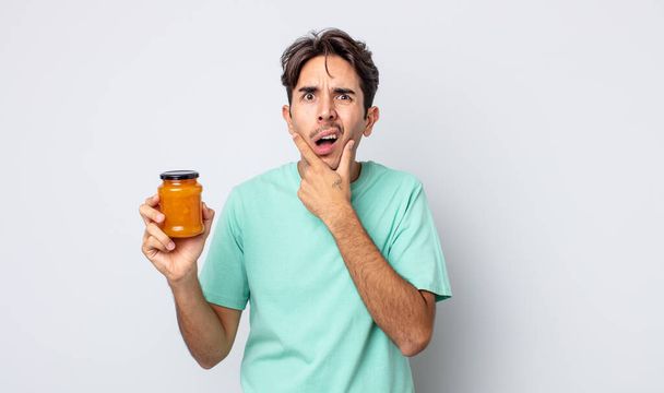 young hispanic man with mouth and eyes wide open and hand on chin. peach jelly concept - Foto, afbeelding