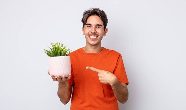 young hispanic man smiling cheerfully, feeling happy and pointing to the side. decorative plant concept - Photo, image