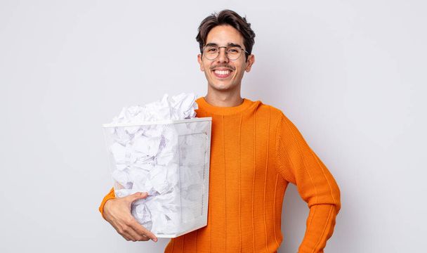 young hispanic man smiling happily with a hand on hip and confident. paper balls trash concept - Foto, Bild