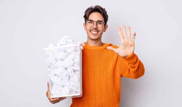 young hispanic man smiling and looking friendly, showing number five. paper balls trash concept - Foto, Bild