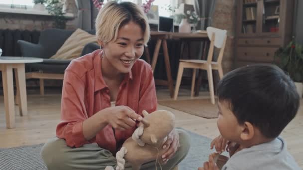 Medium slow-shot di allegra giovane donna asiatica che gioca con il suo intelligente figlio di 3 anni, seduto sul pavimento in un accogliente soggiorno con giocattolo di peluche - Filmati, video