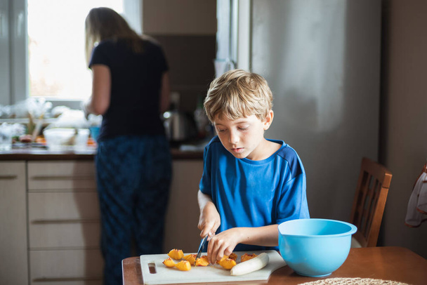 Młody chłopiec w kuchni kroi owoce podczas przygotowywania koktajlu, w tle jego matka - Zdjęcie, obraz
