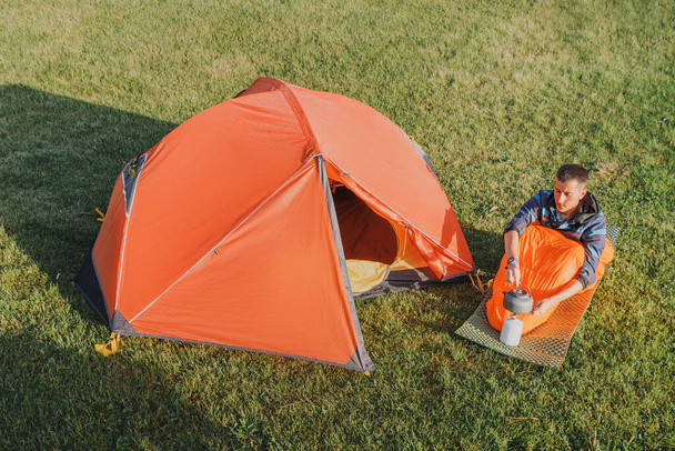 調理キャンプガスストーブやバーナーで男沸騰茶の空中ビュー,早朝に牧草地にオープンオレンジのテントの近く - 写真・画像