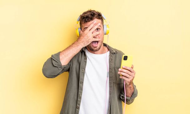 young handsome man looking shocked, scared or terrified, covering face with hand headphones and smartphone concept - Fotografie, Obrázek