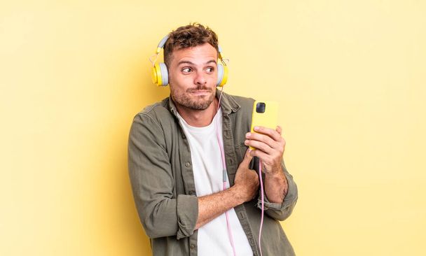 young handsome man shrugging, feeling confused and uncertain headphones and smartphone concept - Foto, imagen