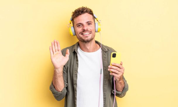 young handsome man smiling happily, waving hand, welcoming and greeting you headphones and smartphone concept - Фото, изображение