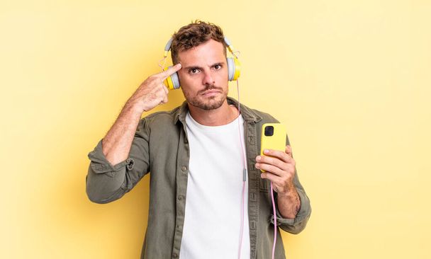 young handsome man feeling confused and puzzled, showing you are insane headphones and smartphone concept - Foto, imagen