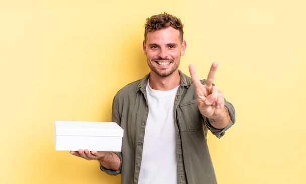young handsome man smiling and looking happy, gesturing victory or peace. white box concept - Фото, зображення