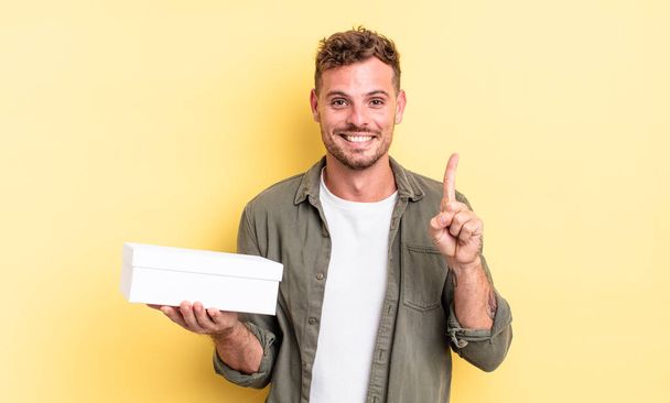 young handsome man smiling and looking friendly, showing number one. white box concept - Foto, Imagen