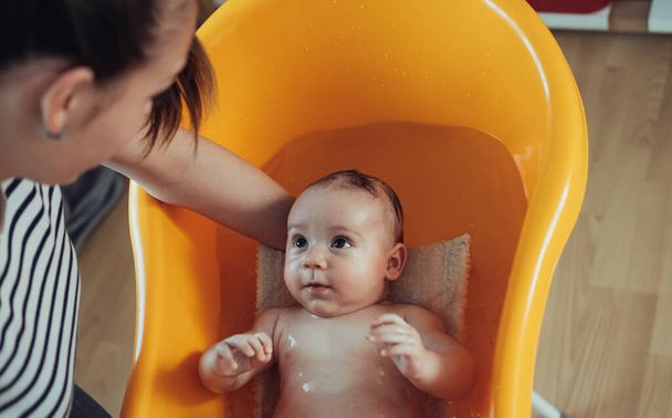 Μη αναγνωρίσιμη μητέρα πλένει το αγοράκι της στο σπίτι. Μια ανώνυμη μητέρα που κάνει μπάνιο ένα νεογέννητο μωρό σε πορτοκαλί μπανιέρα στο σπίτι.. - Φωτογραφία, εικόνα
