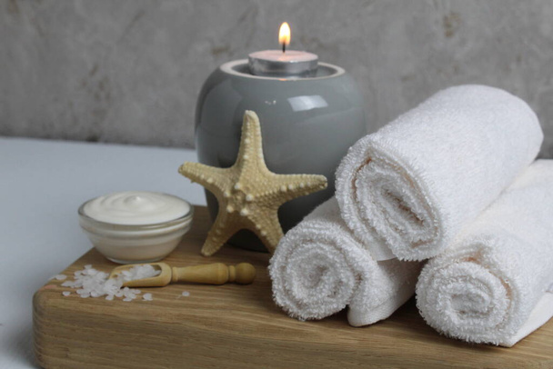 Salz für Bad Massage Peeling Wellness Entspannungsmassage. Körperpflege zu Hause. Schönheit. Weiße Handtücher Seesterne Salzkerzen auf einem hölzernen Tablett auf einem grauen Hintergrund Seitenansicht. - Foto, Bild