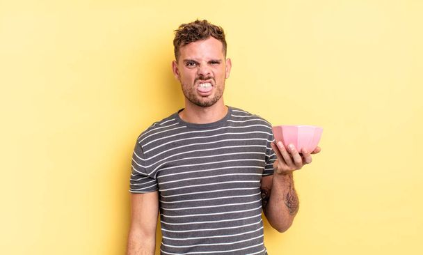 young handsome man feeling disgusted and irritated and tongue out empty bowl concept - Foto, imagen