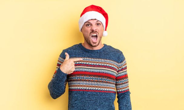 young handsome man looking shocked and surprised with mouth wide open, pointing to self. christmas concept - Foto, afbeelding