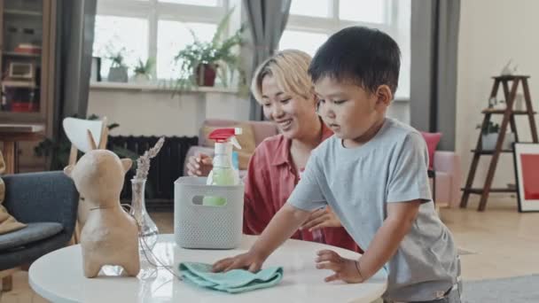 Keskipitkä hidas laukaus söpö pieni aasialainen poika auttaa äitiään tekemään kotitöitä, pyyhkimällä sohvapöytä puhtaammalla - Materiaali, video