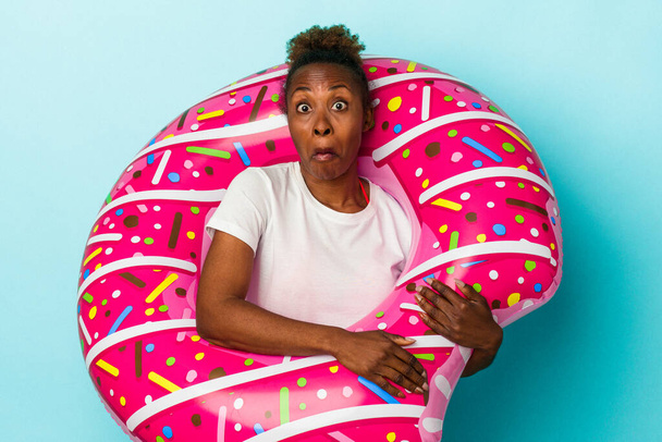 Young african american woman with inflatable donut isolated on blue background shrugs shoulders and open eyes confused. - Photo, Image