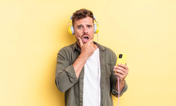 young handsome man with mouth and eyes wide open and hand on chin headphones and smartphone concept - Фото, изображение