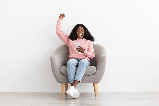 Συναισθηματική μαύρη γυναίκα που παίζει στο διαδίκτυο, χρησιμοποιώντας κινητό τηλέφωνο στο σπίτι - Φωτογραφία, εικόνα
