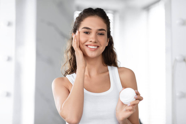 Headshot di gioiosa femmina idratante viso applicando crema in bagno - Foto, immagini