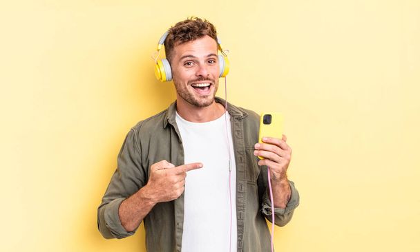 young handsome man looking excited and surprised pointing to the side headphones and smartphone concept - Φωτογραφία, εικόνα