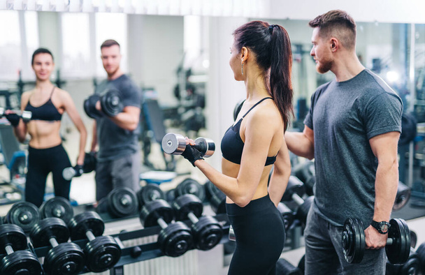Trainer and client looking at the mirror while doing workout at gym with dumbbell. Young brunette with personal fitness trainer. Fitness concept. Closeup. Selective focus - Foto, Bild
