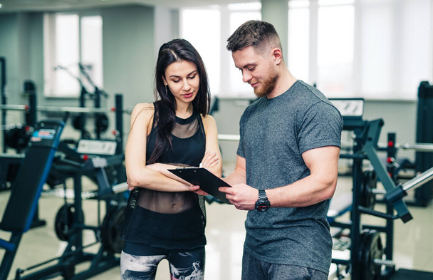 Trainer and client discussing her progress at the gym. Gym instructor writing on clipboard. Stock photo - Фото, изображение