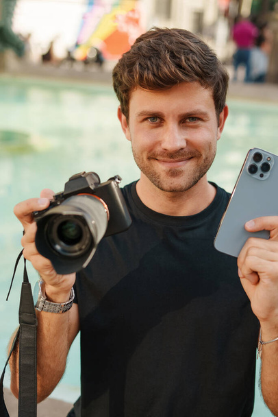 Positivo giovane uomo che tiene smartphone e fotocamere in mano e sorride - Foto, immagini