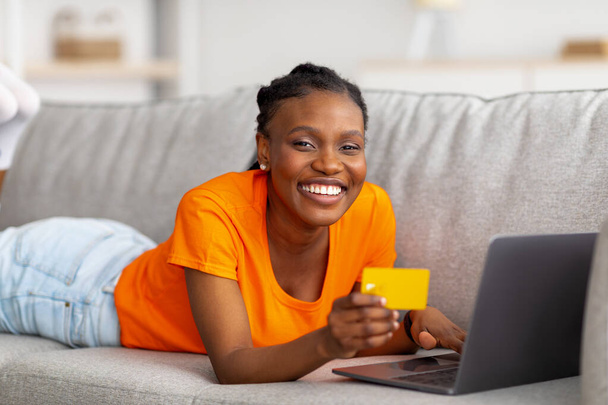 Радісна африканська жінка, яка займається магазином он - лайн за допомогою ноутбука і кредитної картки, лежить вдома на дивані. - Фото, зображення