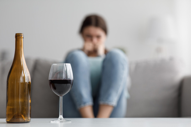 Λυπηρό καυκάσιος χιλιετή γυναίκα κάθεται στον καναπέ που πάσχουν από άγχος και προβλήματα, εστίαση στο μπουκάλι και ένα ποτήρι κρασί - Φωτογραφία, εικόνα