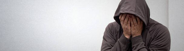 Das Porträt eines jungen erwachsenen Mannes mit grauer Kapuze bedeckte sein Gesicht mit den Händen. Das Konzept von Sucht und Alkoholismus, Depression und Hoffnungslosigkeit - Foto, Bild