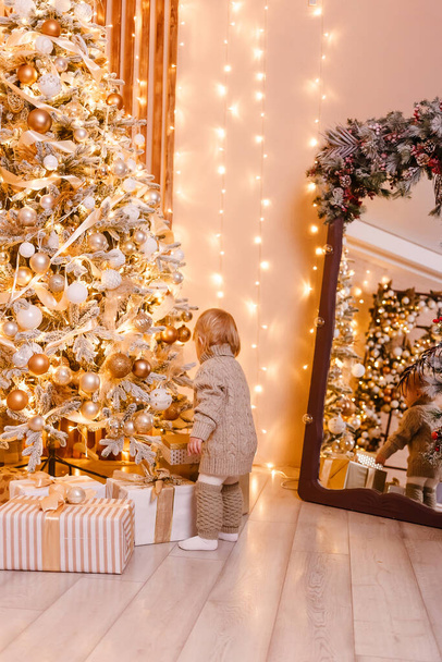 Örgü kazak giyen iki yaşındaki bir bebek daha büyük bir Noel ağacını süslüyor. Sırtı kameraya dönük kız. - Fotoğraf, Görsel