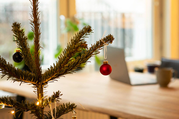 Weihnachtsbaum mit Dekoration. Im Hintergrund, unscharf, steht ein Laptop auf einem Holztisch. Konzept, von zu Hause aus zu arbeiten oder in den Ferien fern zu arbeiten. Foto aus Schweden. - Foto, Bild