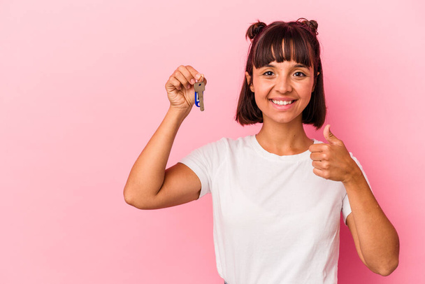 Junge Frau mit gemischter Rasse hält einen Schlüssel isoliert auf rosa Hintergrund lächelnd und hebt den Daumen - Foto, Bild