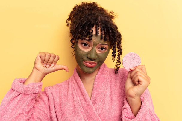 Jovem mulher de raça mista vestindo um roupão de banho segurando uma esponja removedor de maquiagem isolada no fundo amarelo se sente orgulhosa e auto-confiante, exemplo a seguir. - Foto, Imagem