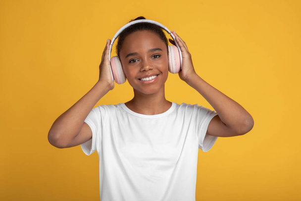 Cheerful pretty teen afro american girl in white t-shirt in earphones listen music, isolated on yellow background - Photo, Image