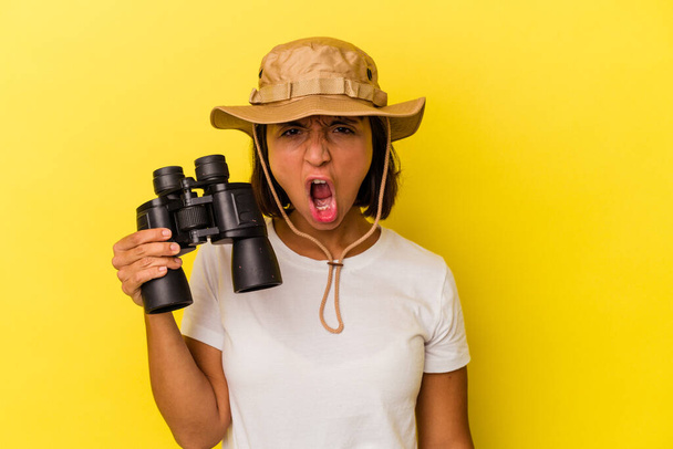 Young mixed race explorer woman holding binoculars isolated on yellow background screaming very angry and aggressive. - Photo, Image
