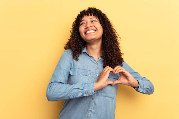 Mujer joven de raza mixta aislada sobre fondo amarillo sonriendo y mostrando una forma de corazón con las manos. - Foto, imagen