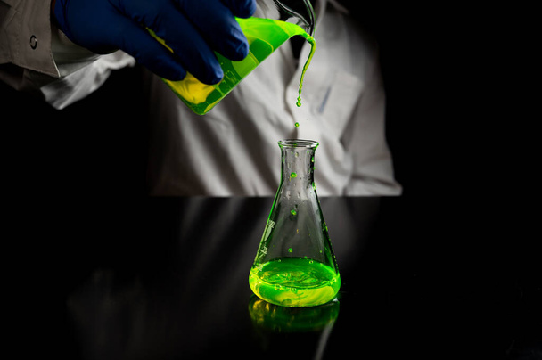 Ein Forscher experimentiert mit grünen fluoreszierenden Tröpfchen in einem Glaskolben in einem dunklen biomedizinischen Labor für die Entwicklung der Gesundheitsmedizin. Kopierraum schwarzer Hintergrund - Foto, Bild