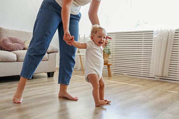 First steps of a little girl. A blond toddler learning to walk at home with the help of loving mother. Close up, copy space for text, background. - Valokuva, kuva