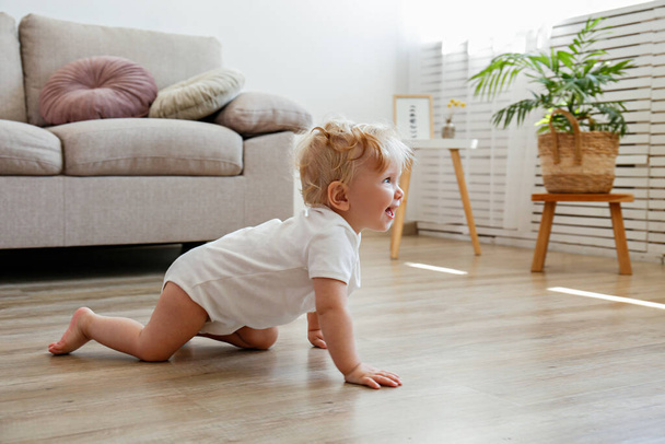 One year old child learning to crawl on the wooden floor of living room. Interior background. Adorable blonde little girl in pink crawlers at home. Close up, copy space for text. - Фото, зображення