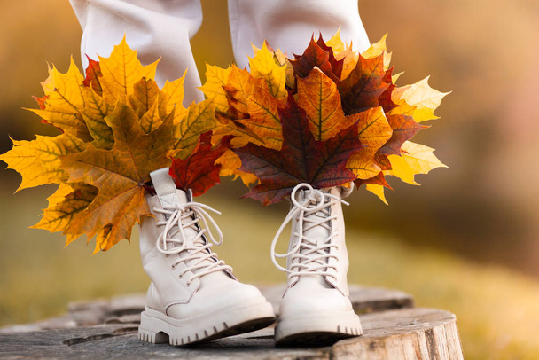 Nahaufnahme beiger Stiefel an weiblichen Beinen mit bunten Ahornblättern. Herbststimmung. Stilvolle Schuhe, Herbstmode, Herbstverkauf. Selektiver Fokus. - Foto, Bild