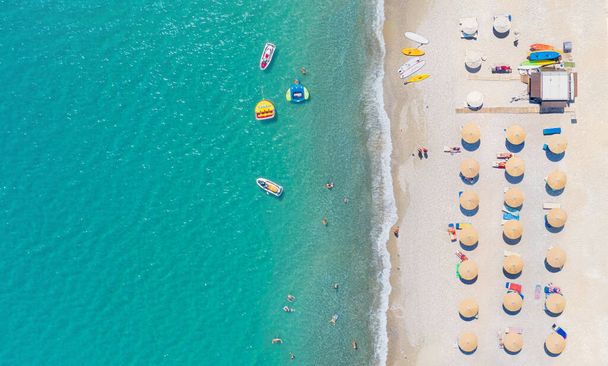 Песчаный пляж с шезлонгами, зонтиками и оборудованием для водных видов спорта, люди плавают и принимают солнечные ванны. Воздушная панорама - Фото, изображение
