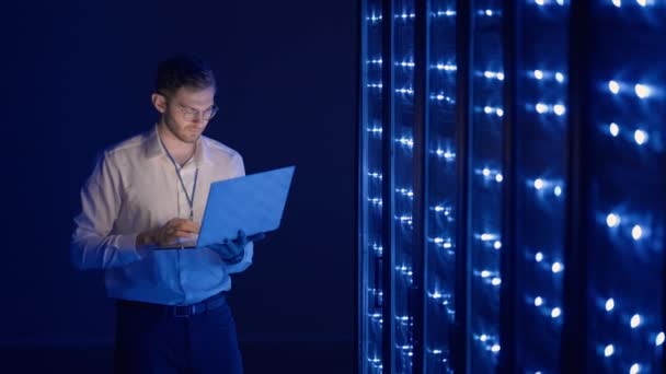 Mężczyzna inżynier sieci robi kontrolę systemu stojąc w serwerowni z laptopem. W centrum danych mężczyźni serwer specjaliści sprawdzanie systemu pracy i sprzętu szafy serwerowe rack - Materiał filmowy, wideo