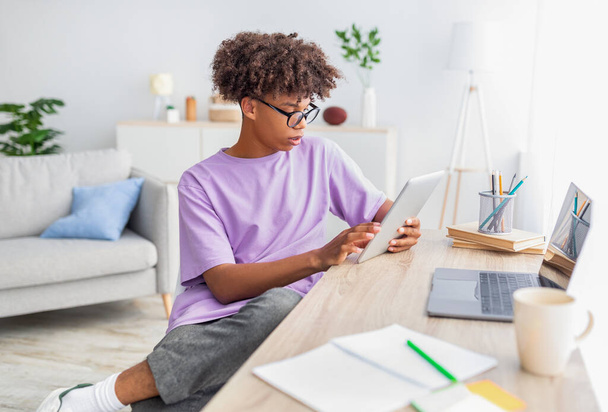 Adolescente negro inteligente que usa tableta y computadora portátil para estudios en línea en casa. Concepto de escolaridad doméstica - Foto, imagen