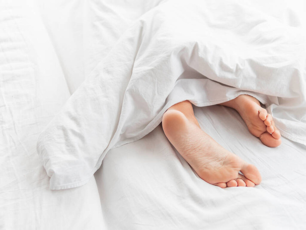 View on bare feet under blanket. Bedroom lit with morning light. Woman sleeps with comfort. White crumpled linen. - Photo, Image